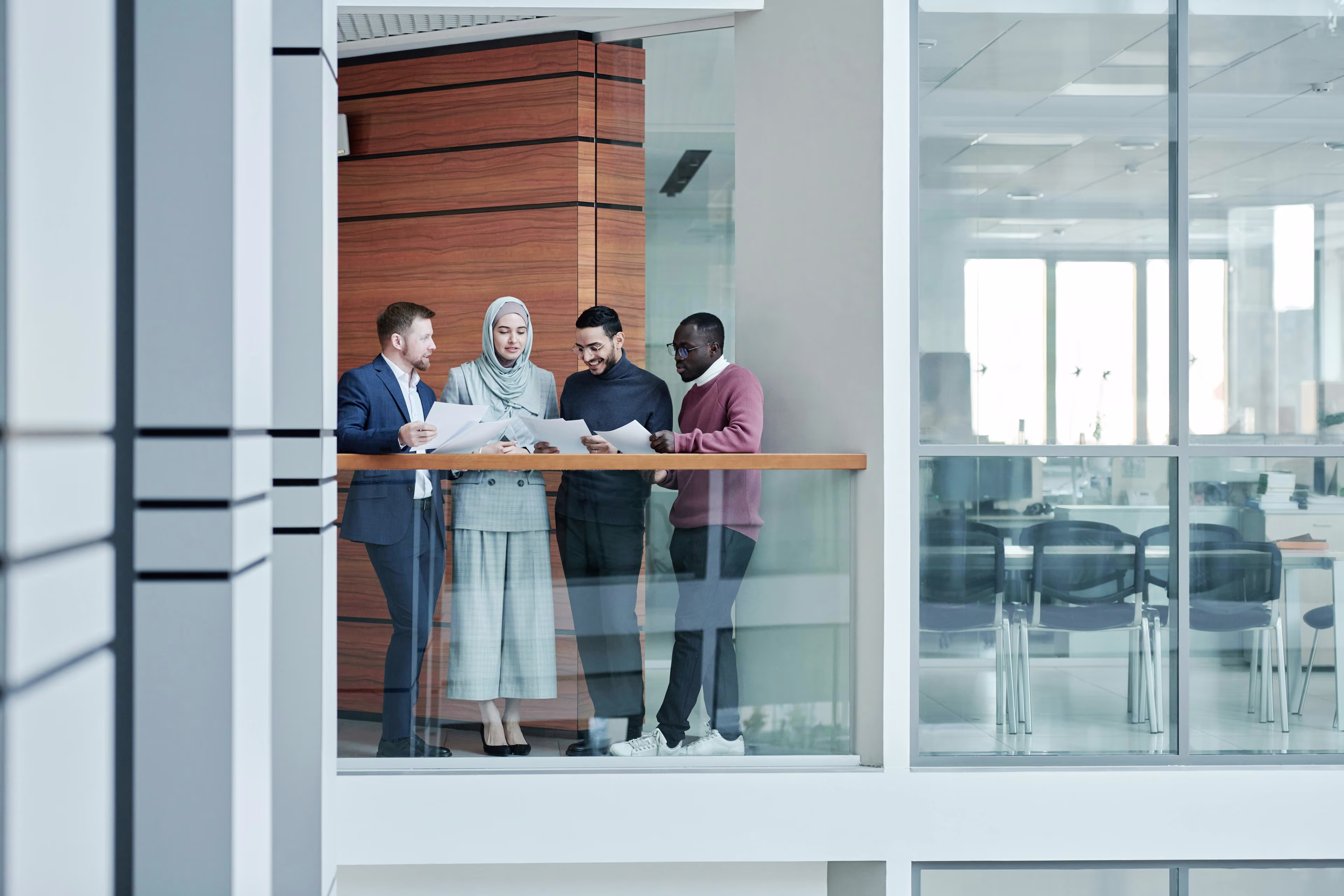 Diverse group of four business professionals standing in a modern office corridor reviewing documents together.