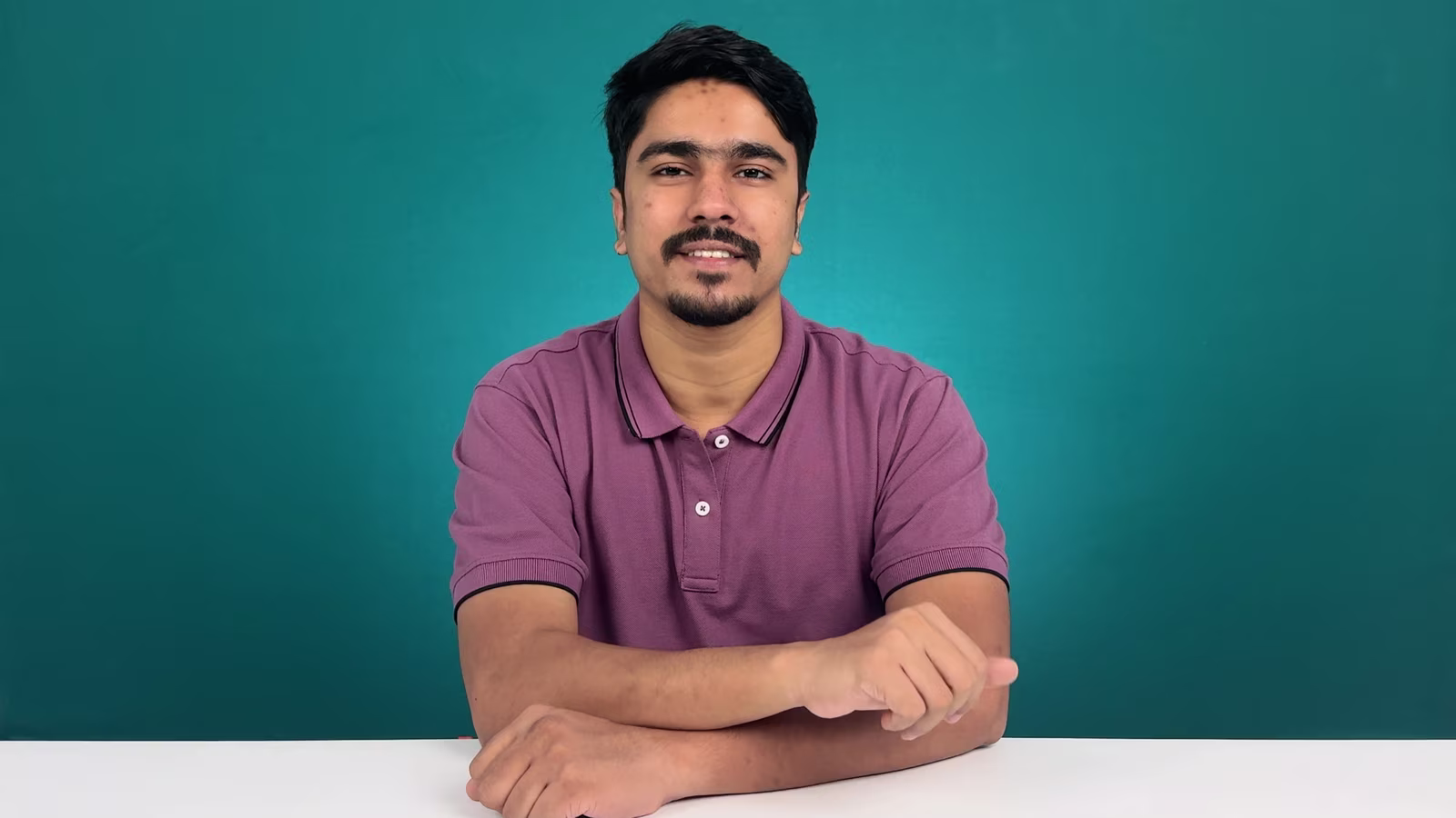 Man sitting at a white table against a teal background, wearing a mauve polo shirt and looking toward the camera with a neutral expression.
